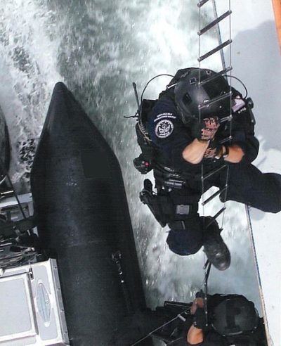 Members of a United States Coast Guard Maritime Response Team descend towards a target while waves crash onto the ship's deck.