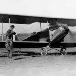 Charles Lindbergh adjusts his parachute in front of Sergeant Bell’s experimental biplane on Lambert Field, St. Louis, Missouri, in the mid-to-late 1920s, while Bell waits in the plane.