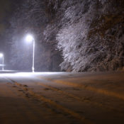 A snow-covered road at night. It cuts through a wooded area, and street lights glow brightly.
