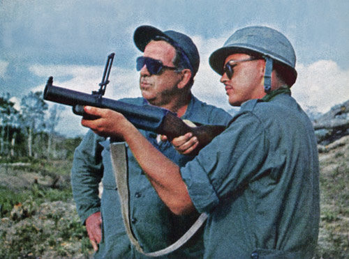 A soldier holds a grenade launcher while his father looks on.