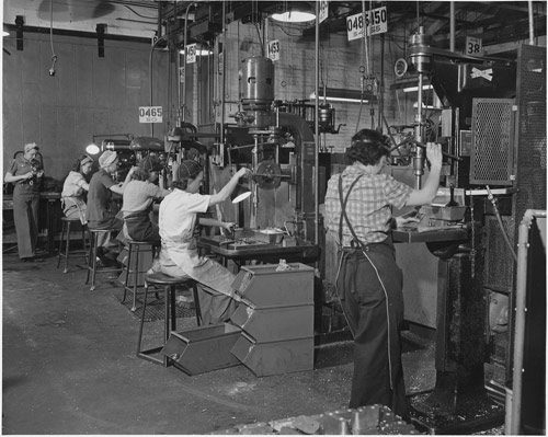 A team of women workers operate machinery in an airplane factory. Due to the dangerous nature of the job, the workers are wearing pants instead of dresses.
