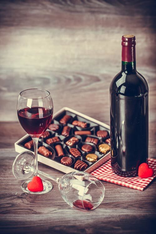 A bottle of red wine, a filled wine glass, and an open box of chocolates are on a wooden table.