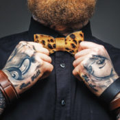 A bearded man with wrist tattoos fixes his bowtie.