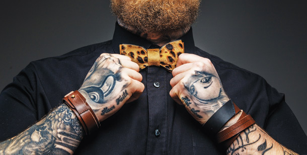 A bearded man with wrist tattoos fixes his bowtie.