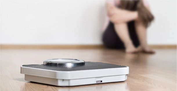 A sullen woman sits next to a weight scale.