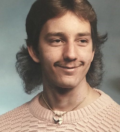 Man with a thin mustache and a blond mullet poses for portrait.