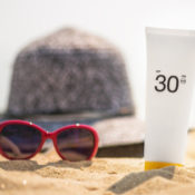 A bottle of sunscreen, a pair of sunglasses, and a hat in the sand on a bright, summer day.
