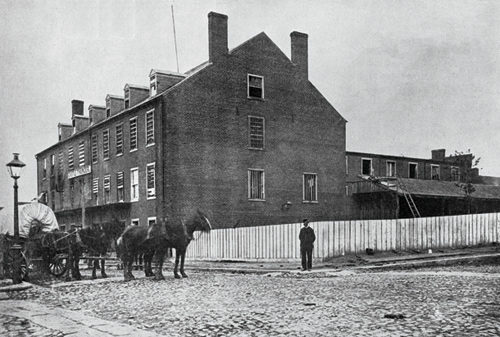 The outside of a tabacco warehouse that was converted into a prison during the Civil War. Horses pull a covered wagon in front of the warehouse.