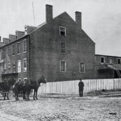 The outside of a tabacco warehouse that was converted into a prison during the Civil War. Horses pull a covered wagon in front of the warehouse.