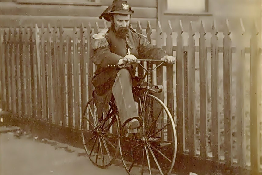 Bearded man wearing a miltary uniform and hat similar to Napoleon rides a bicycle.