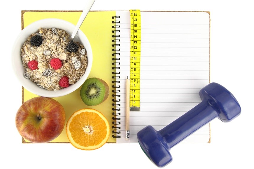 A collection of healthy living items, including a bowl of oatmeal, fruit, and a small weight, arranged on a notepad.