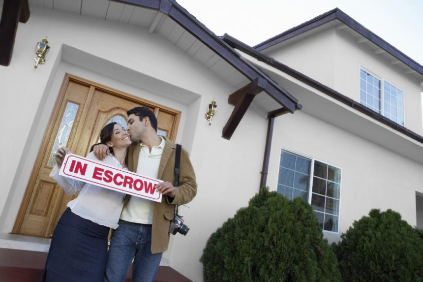 Couple standing in front of a house with a sign that reads "In Escrow"