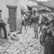 A prisoner lays against a wall as Mexican soldiers point their guns towards him