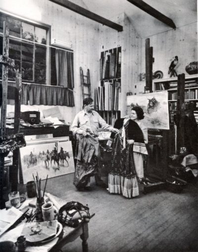 Artist Harold von Shmidt and his wife examine vintage American West clothing in his art studio. Examples of his work, featuring cowboys and other symbols of the Old West, hang on easels.