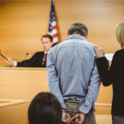 A handcuff man stands in front of a judge while his lawyer places her hand on his shoulder