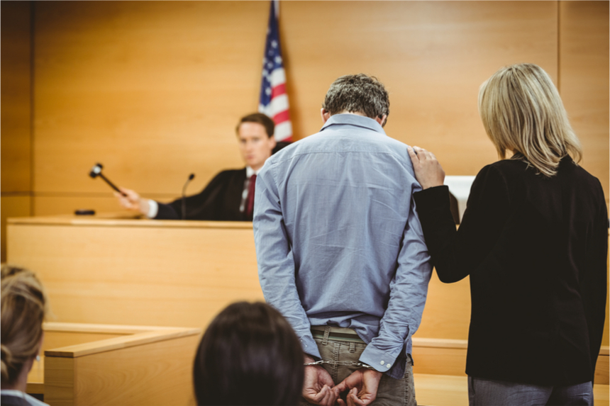 A handcuff man stands in front of a judge while his lawyer places her hand on his shoulder