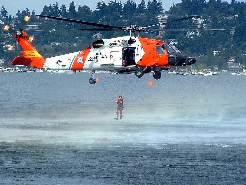 A member of the United States Coast Guard demonstrates a rescue operation by descending from a helicopter midflight over water.
