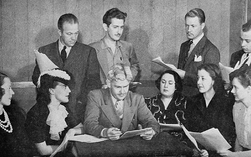 Soap opera cast members crowd around a table during a script reading.
