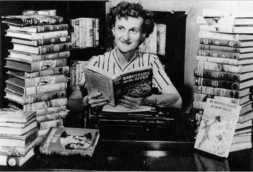 Nancy Drew author Carolyn Keene (real name, Mildred Augustine Writ Benson) sits at a desk with a collection of her books.