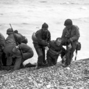 Soldiers landing on the Normandy beaches during the D-Day invasions; a pair of soldiers help their wounded mate onto the rocky shore.