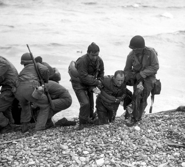 Soldiers landing on the Normandy beaches during the D-Day invasions; a pair of soldiers help their wounded mate onto the rocky shore.