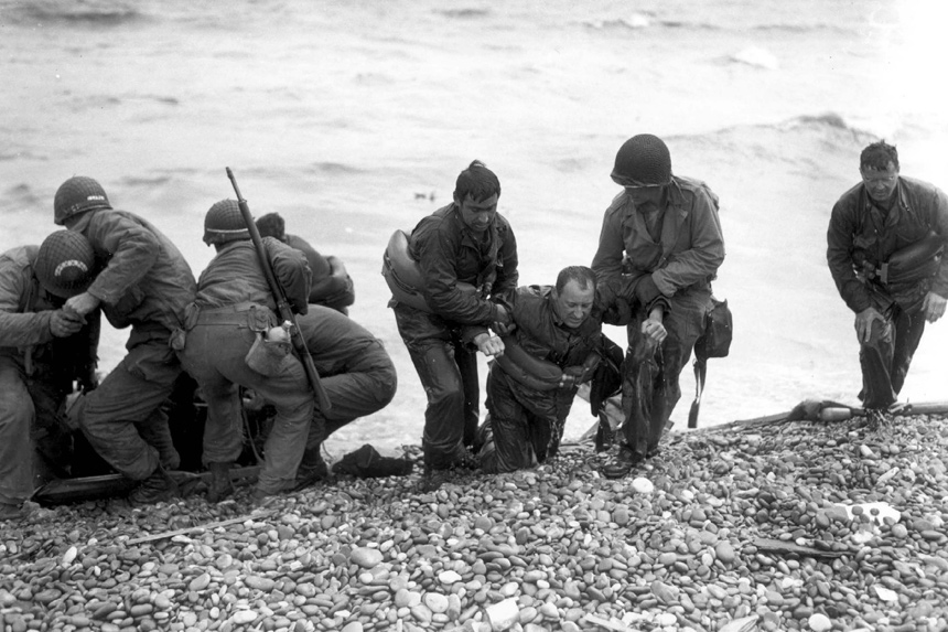 Soldiers landing on the Normandy beaches during the D-Day invasions; a pair of soldiers help their wounded mate onto the rocky shore.