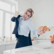 Woman preparing for a work day by putting on a jacket, carrying a cup of coffee, and grabbing a pastry off her kitchen counter.