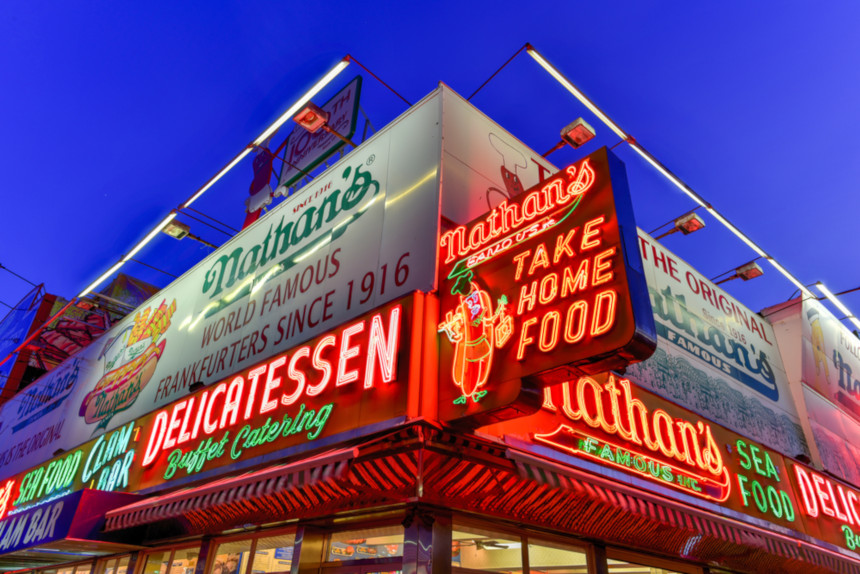 The modern Nathan's Famous Hotdogs restaurant at Coney Island, at night. The building's facade is covered with neon signs.