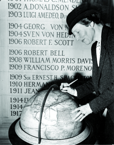 American explorer Louise Arner Boyd signing the Fliers and Explorers Globe, as it rests in front of a wall featuring the names of other signatories.