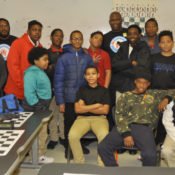 Chess teacher Orrin Hudson poses with his students in a classroom, flanked by a chess table.