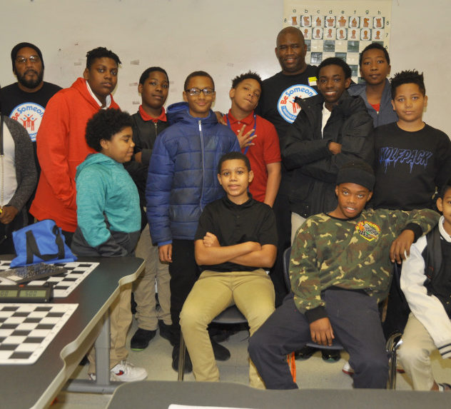 Chess teacher Orrin Hudson poses with his students in a classroom, flanked by a chess table.
