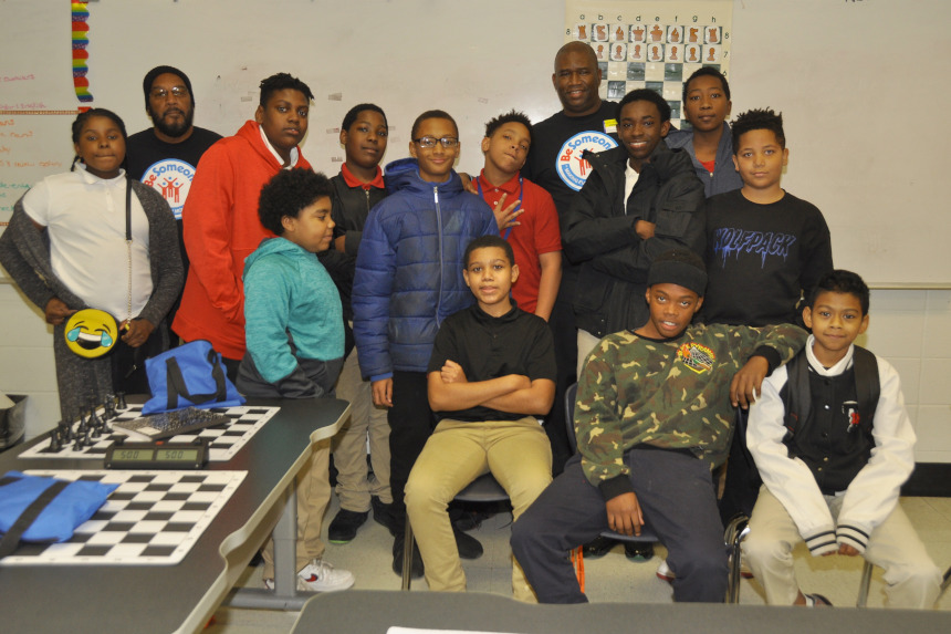 Chess teacher Orrin Hudson poses with his students in a classroom, flanked by a chess table.
