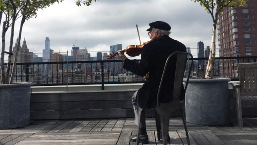 A scene from the documentary, "Fiddler: A Miracle of Miracles" that shows an older man playing the fiddle in a city park.