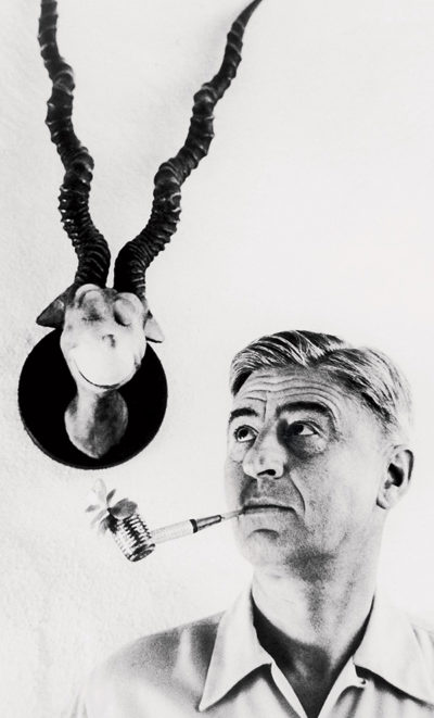 Theodore Geisel — also known as "Dr. Seuss" — looks up at a bust of one of his characters mounted on a wall while holding a flower-filled pipe in his mouth.