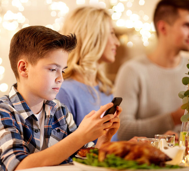 A young boy looking at his smartphone while he and his family are having dinner.