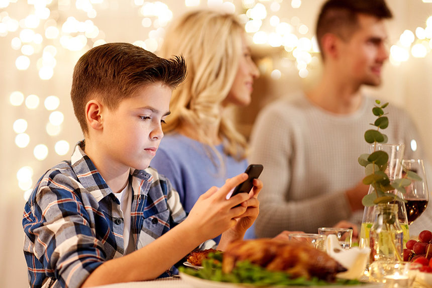 A young boy looking at his smartphone while he and his family are having dinner.
