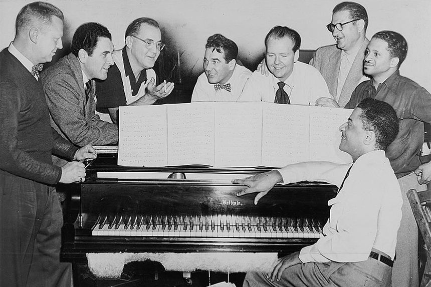 Benny Goodman (third from left) with some of his former musicians, seated around piano left to right: Vernon Brown, George Auld, Gene Krupa, Clint Neagley, Ziggy Elman, Israel Crosby and Teddy Wilson (at piano).