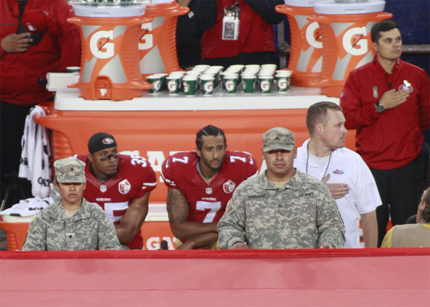 Colin Kaepernick and Eric Reid kneel during the national anthem on a football field's sidelines.