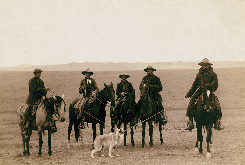 Cowboys on horseback pose with a tied-up wolf.