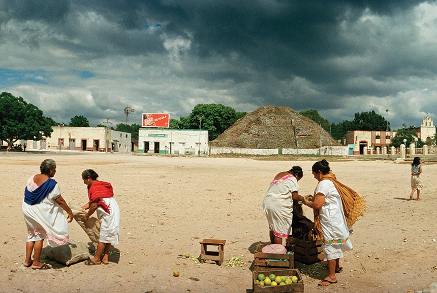 Women pack fruit into their sacks outside a market in Acanceh, Yucatán, Mexico. An ancient mound can be seen, nearby.