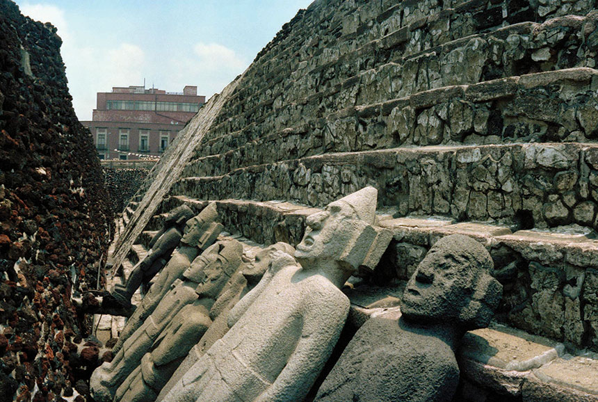 Ancient statues rest against old Mayan stairs near modern buildings, in Mexico City.