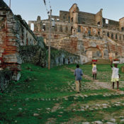 Young women carrying baskets of food and laundry past ancient ruins.