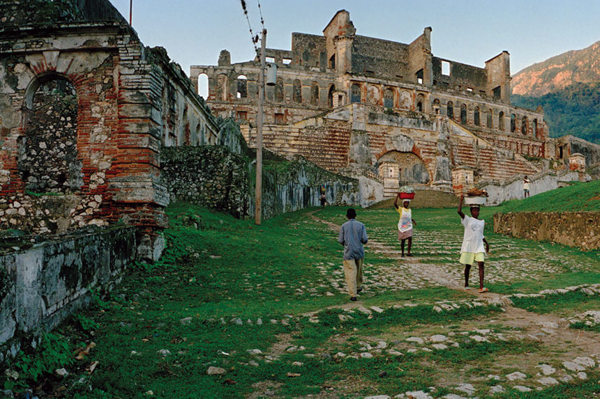 Young women carrying baskets of food and laundry past ancient ruins.