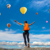 Hiker on a mountain raises their hands to greet a group of hot air balloons