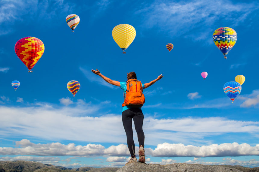 Hiker on a mountain raises their hands to greet a group of hot air balloons