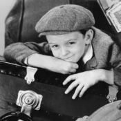 kid dressed in early 20th-century clothes sits on a travel suitcase