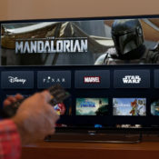 Man holding a remote control in front of a television that has Disney+ displayed. The Mandalorian is being advertised on the app.