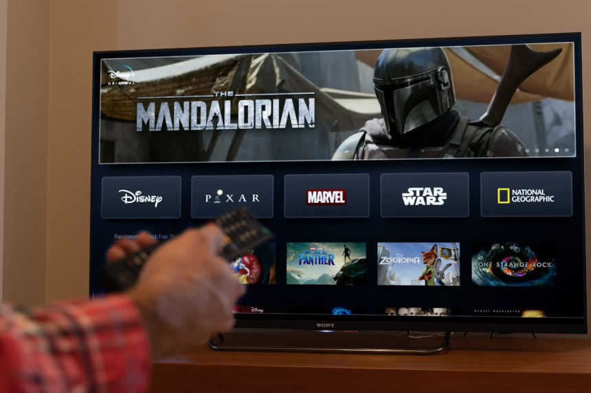 Man holding a remote control in front of a television that has Disney+ displayed. The Mandalorian is being advertised on the app.
