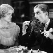 1920s image of a man eating a large turkey leg while a woman looks at him disapprovingly