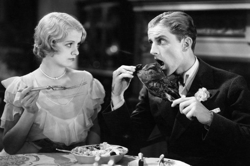 1920s image of a man eating a large turkey leg while a woman looks at him disapprovingly
